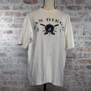 May include: White short-sleeved t-shirt with a graphic print of a compass, sailboat, and oars. The text "SAN DIEGO" is printed above the graphic and "NAUTICA" is printed below the graphic.