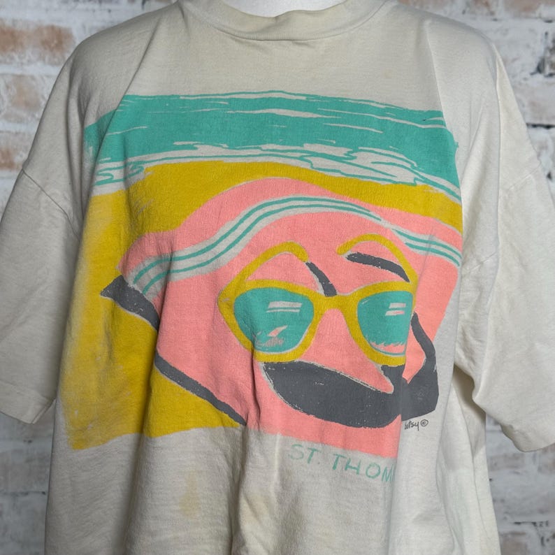 This image showcases the upper front side, the shoulders, the sleeves and the middle front side of a true vintage 1980s salvage condition thrashed Belton Tough Tee women’s 100% preshrunk cotton St. Thomas single stitch graphic t-shirt in a size XL modeled on a dress form in front of a brick backdrop.