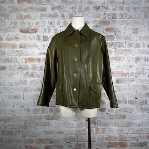 May include: Olive green leather jacket with a classic collar, button-up front, and two front pockets. The jacket is displayed on a mannequin against a brick wall background. The jacket has long sleeves and a tailored fit.