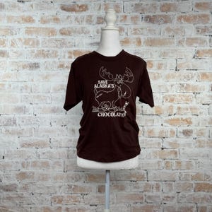 May include: A dark brown t-shirt with a white graphic of a moose and the text "Save Alaska's Chocolate". The shirt is displayed on a mannequin against a brick wall background.