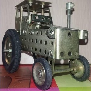 Peut inclure: Un tracteur jouet de couleur argentée, construit à partir de pièces métalliques, d'écrous et de boulons. Le tracteur comprend une cabine, deux grandes roues arrière et deux petites roues avant avec des pneus en caoutchouc noirs. Le modèle a une cheminée.