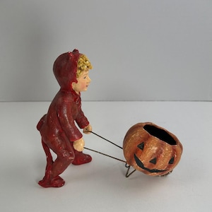 May include: A Halloween figurine depicts a child in a red devil costume pulling a jack-o'-lantern. The jack-o'-lantern is orange with a carved face and sits on a metal frame. The child has blonde hair and a painted face.