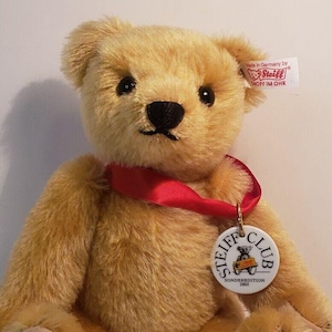 Puede incluir: Un oso de peluche beige Steiff con una cinta roja y una medalla del Steiff Club. La medalla es blanca con un logotipo Steiff rojo y negro y el texto "Sonderedition 2002". La pata del oso tiene el texto "1993 06887" impreso en ella.