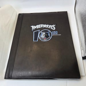 May include: A dark brown leather binder with the embroidered text "TIMBERWOLVES 10TH ANNIVERSARY". The logo features a wolf with a blue and white color scheme. The binder is closed and has a smooth texture.