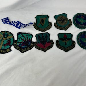 May include: A collection of vintage military patches and a blue ribbon with white text. The patches feature various shield and circular designs with green, gold, red, and blue colors. Some patches include text such as "AIR FORCE" and "AIR TRAINING COMMAND".
