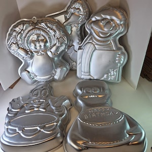 Puede incluir: Una colección de moldes para pasteles de metal vintage en varias formas de personajes. Los moldes incluyen diseños de una muñeca, Big Bird, Ernie y un personaje sosteniendo un pastel con la inscripción "Happy Birthday". Los moldes son de color plateado.