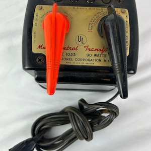 May include: A black Lionel model train transformer with a gold faceplate and red and black control levers. The transformer has a power cord and plug. The faceplate has text that reads "Lionel Corporation, N.Y." and "Made in U.S. of America."