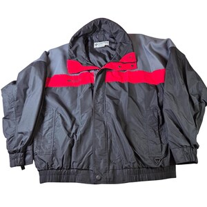 May include: A black, gray, and red Columbia jacket. The jacket has a high collar and a red stripe across the chest. The Columbia logo is visible on the chest and collar. The jacket has a button closure and two pockets.