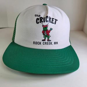 Może przedstawiać: Biało-zielona czapka typu trucker z napisem "the CRICKET" i postacią z kreskówki przedstawiającą świerszcza w czerwonej kurtce. Świerszcz trzyma zielony worek z pieniędzmi. Poniżej znajduje się napis "ROCK CREEK, MN.".