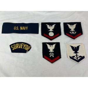 Op de afbeelding: Een verzameling Amerikaanse marine-patches en insignes. Bevat een donkerblauwe patch met "U.S. NAVY" in gouden letters, een "SURVEYOR" patch en verschillende patches met adelaars en diverse symbolen.