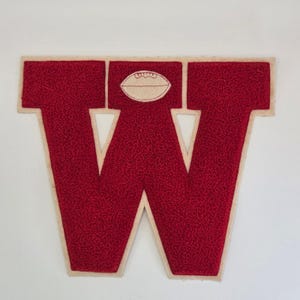 May include: A red felt letter "W" with a football graphic. The letter has a textured surface and a cream-colored border. The football is cream-colored with brown stitching. The letter is likely a patch for a varsity jacket or sports apparel.