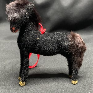 May include: A small, black, fuzzy horse figurine with a textured, flocked surface. The horse has a dark mane and tail, gold-colored hooves, and a thin red ribbon tied around its neck. The figure stands in a profile position on four legs.