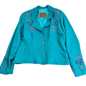 May include: A turquoise blazer featuring a notched lapel, three silver-toned buttons, and long sleeves. The garment is decorated with embroidered floral patterns in pink, purple, and yellow on the chest and cuffs. A brown brand label is visible.