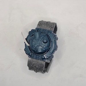 Puede incluir: Un reloj de pulsera de juguete azul oscuro y gris con una esfera circular y una correa texturizada. La esfera del reloj tiene un diseño mecánico detallado con un botón central. La correa es gris y presenta un patrón texturizado.