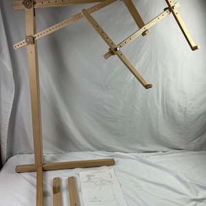 Puede incluir: Un telar o soporte de bordado de madera ajustable. El soporte está hecho de madera de color claro y presenta una base en forma de cruz, brazos ajustables y un papel con instrucciones. Incluye dos piezas de madera adicionales.