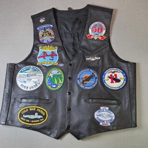 May include: Black leather vest with various embroidered patches. The patches display text and images related to submarines, including "Cold War Patrol" and "United States Submarine Veterans". The vest has a button-up front and two pockets.