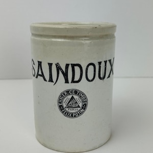 May include: Off-white ceramic jar with the word "SAINDOUX" in large black letters. Below the text is a circular emblem with text and a triangle. The jar has a cylindrical shape with a slightly raised rim.