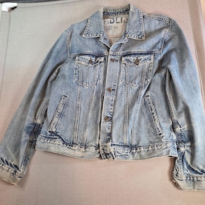 May include: Pale blue denim jacket with a traditional collar, button closure, and several pockets. The jacket has a faded wash and a worn look. The brand name "ADLIS" is visible on the collar.