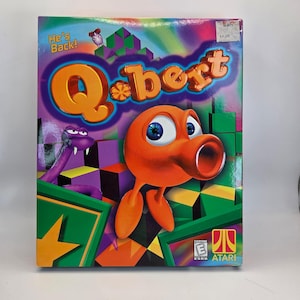 May include: A colorful Q*bert video game box featuring the orange Q*bert character with blue eyes and a red mouth. The box has a purple background with the words "He's Back!" and the game's title in orange. The box also features a purple snake.