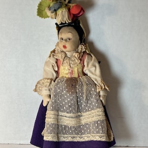Puede incluir: Una muñeca vintage vestida con atuendo tradicional. La muñeca tiene una cara pintada, un tocado floral y viste una blusa color crema con adornos de encaje. La falda de la muñeca es morada con una superposición de encaje blanco y tiene zapatos morados a juego.