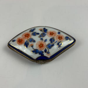 Könnte beinhalten: Dekorative, fächerförmige Schmuckschatulle mit weißer Porzellanoberseite. Die Oberseite zeigt ein Blumendesign mit orangefarbenen Blüten, blauen Blättern und goldenen Akzenten. Die Schatulle hat einen blauen Rand und einen goldfarbenen Metallrahmen.