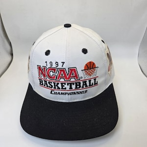May include: White and black baseball cap with the text "1997 NCAA Basketball Championship". The cap features a basketball and net graphic. The brim is black, and the top is white.