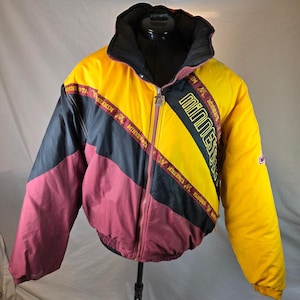 Puede incluir: Una chaqueta con cremallera que presenta un diseño de bloques de color amarillo, negro y granate. La chaqueta muestra la palabra Minnesota en texto amarillo en el pecho e incluye un patrón de texto Minnesota repetido en franjas diagonales.