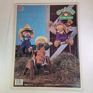 May include: A Cabbage Patch Kids 25-piece jigsaw puzzle showing three dolls in overalls and hats, set against a barn backdrop. The puzzle is designed for ages 3 to 7 and includes the MB logo.