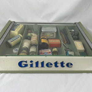 May include: Vintage Gillette display case with a variety of grooming products. The case is light green with a glass top and the word "Gillette" in blue. Inside are shaving brushes, bottles, tins, and other grooming accessories.