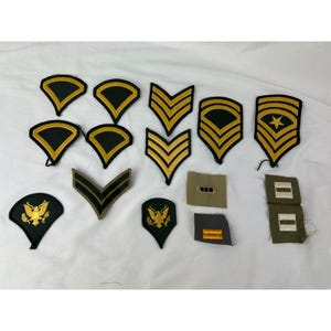 Puede incluir: Una colección de parches e insignias militares vintage. Los parches presentan varias formas y diseños, incluyendo galones, estrellas y emblemas de águilas. Los parches son en tonos de verde, negro y dorado.