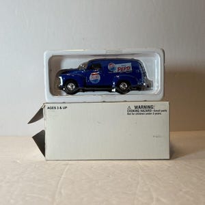 May include: A die-cast blue Pepsi delivery truck toy in a clear plastic case, sitting on top of a white box. The truck has the Pepsi logo on the side and top. The box has the text "AGES 3 & UP" and a choking hazard warning.