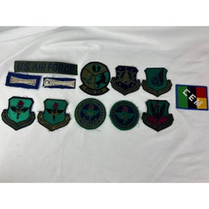May include: A collection of military patches and insignia, including a green and black "U.S. AIR FORCE" patch, several shield-shaped patches with various emblems, and a rectangular patch with the letters "CEM". The patches are in various colors.