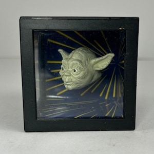May include: A small, light green Yoda figure in a black display case. The figure has large ears and green eyes. The background is dark blue with yellow lines radiating outwards. The display case is square.