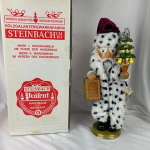 May include: A Steinbach Santa Claus nutcracker figurine with a white and black spotted coat, holding a small Christmas tree and a book. The figurine is on a green base and comes with its original box.