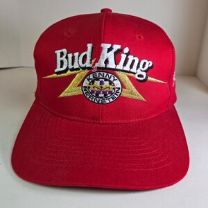 May include: A red baseball cap with the text "Bud King" embroidered in white. The cap features a gold and white emblem with the words "Kenny Bernstein" and a purple and gold design. The cap has a curved brim and a button on top.