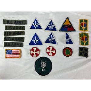 May include: A collection of military patches and insignia, including U.S. Army name tapes, American flag patch, blue triangle patches with a white emblem, and various unit patches with unique designs and colors. The patches are arranged on a white surface.