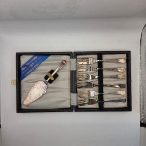 May include: A vintage silver serving set in a black box. The set includes a cake server with an ornate handle and six forks. The box has a blue ribbon with text that reads "Beau Silvermiths Sheffield England".