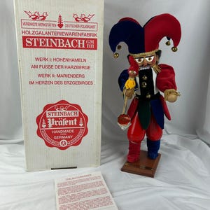 May include: A Steinbach nutcracker figurine in a jester's outfit, with a red, blue, and gold color scheme. The figurine is handmade in Germany and stands on a wooden base. The box next to it has German text.