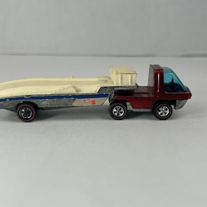 Puede incluir: Un camión de juguete Hot Wheels rojo y azul vintage con un remolque que transporta un barco blanco. El camión tiene un parabrisas azul transparente y ruedas rojas. El remolque es blanquecino con rayas azules y ruedas rojas.