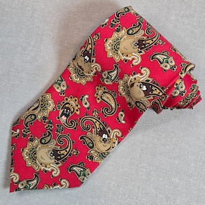 May include: A red necktie with a paisley pattern featuring cartoon character faces in tan, brown, and black. The tie has a classic design, with a pointed end and a folded appearance. The background is a solid light gray.