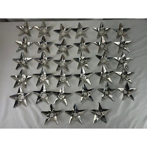 May include: A collection of thirty-one identical silver-toned metal star-shaped ornaments arranged in a grid. Each five-pointed star features a circular cutout in the center and a reflective, polished surface finish.