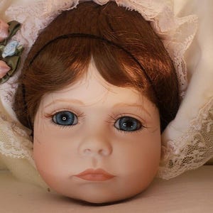 May include: A porcelain doll with brown hair and blue eyes, wearing a white lace bonnet and a pink dress with lace trim.