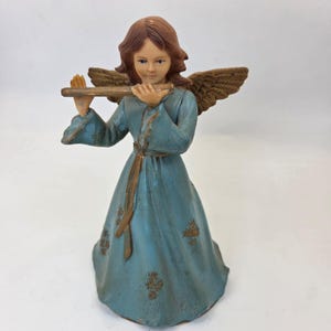 May include: A figurine of an angel with brown hair and textured brown wings, wearing a long blue dress with a waist tie and gold floral accents. The angel is holding a horizontal brown flute-like instrument to its mouth with both hands.