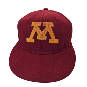 May include: A maroon baseball cap featuring a large, embroidered gold letter M on the front panel. The cap has a structured crown, a flat brim, and small ventilation eyelets stitched into the top panels, all in a matching deep red fabric.
