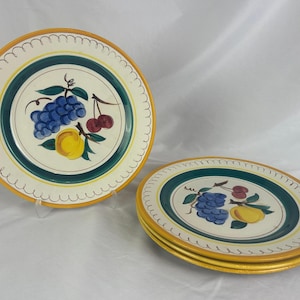 Puede incluir: Un juego de platos decorativos de cerámica con un diseño de frutas. Los platos tienen un centro blanco con un racimo de uvas azules, cerezas rojas y un limón amarillo. Los platos tienen un borde festoneado amarillo y un borde verde y amarillo.