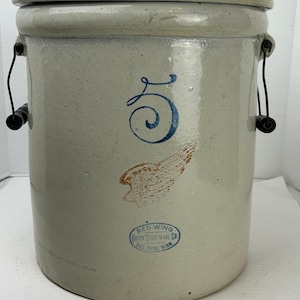 May include: A vintage stoneware crock with a lid. The gray-beige crock features a blue number 5, a brown wing graphic, and the Red Wing Union Stoneware Co. logo. It has black wire handles and a light beige lid.