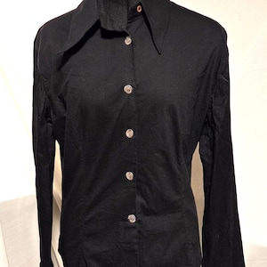 May include: A black button-down shirt with a wide collar and long sleeves. The shirt features decorative silver buttons down the front. The garment is displayed on a black mannequin, showcasing its tailored fit and classic design.