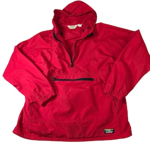 May include: A bright red anorak-style jacket with a hood and a half-zip closure. The jacket has a front pocket with a black zipper and a small logo patch. The sleeves are long, and the jacket appears to be made of a lightweight, water-resistant material.