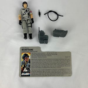 Puede incluir: Una figura de acción de G.I. Joe Mainframe con accesorios, incluyendo una radio, un cable negro y una tarjeta de archivo. La figura lleva un uniforme gris y un casco negro. La tarjeta de archivo tiene el texto "Mainframe Computer Specialist".