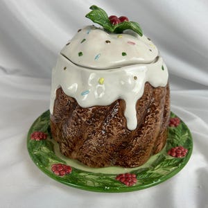May include: A ceramic cake-shaped jar with a lid, decorated with white icing and sprinkles. The jar sits on a green plate adorned with red berries and green leaves. The jar has a brown, textured exterior.